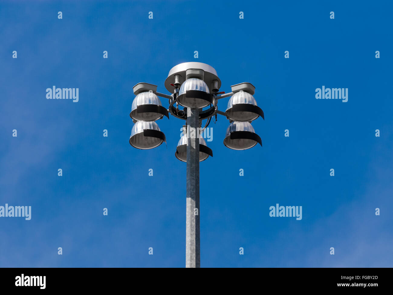 Top of metallic flood lights on metal pole, with six lights pointing down, against blue sky. Stock Photo