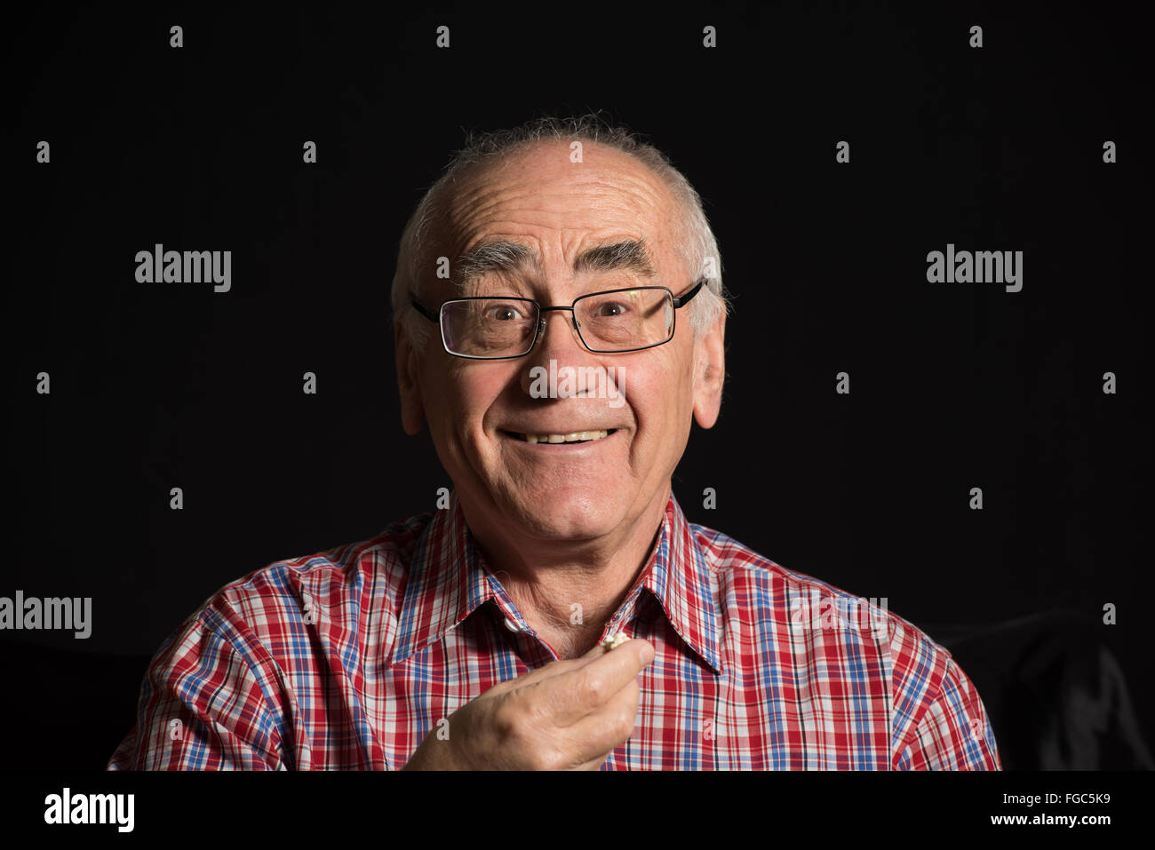old man, wearing checked shirt eats popcorn and watches television