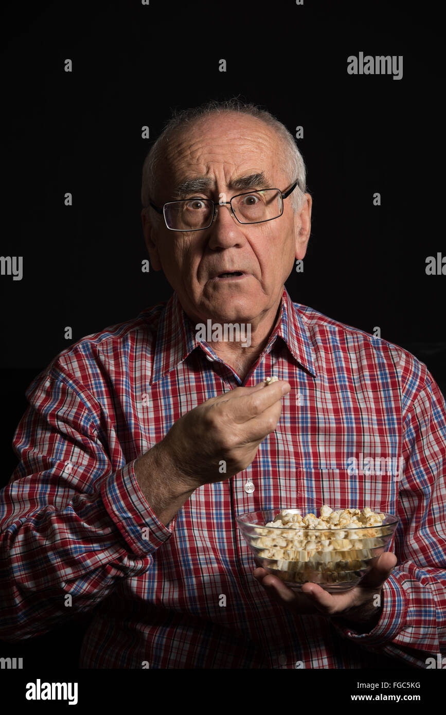 old man, wearing checked shirt eats popcorn and watches television