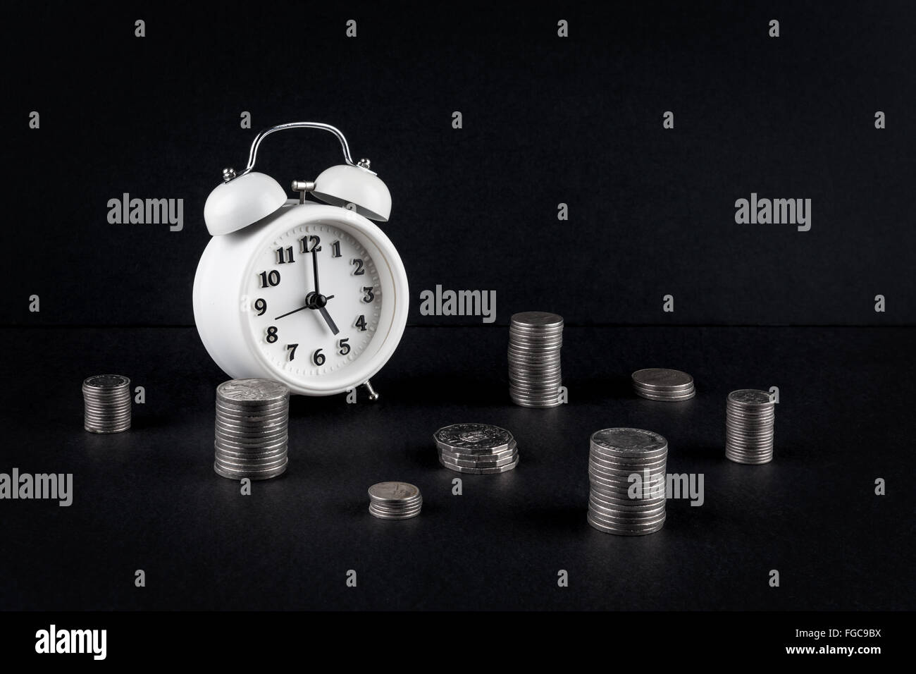 Vintage alarm clock showing 5 o'clock and coin towers on black background. Time is money business concept Stock Photo
