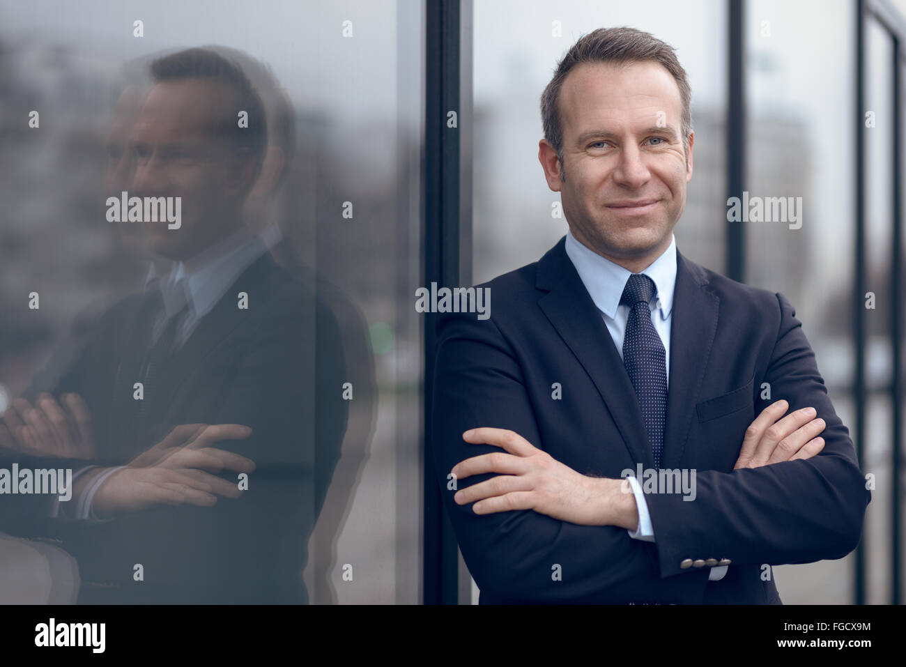 Single confident and handsome male businessman in blue suit and necktie with grin leaning on window outdoors Stock Photo