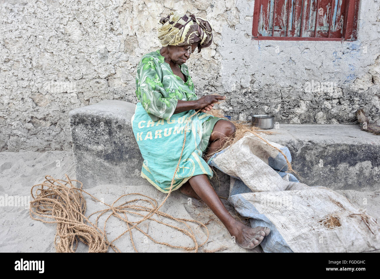 Zanzibari old woman with colourful attire making a rope with coconut fibers outside her home. Stock Photo