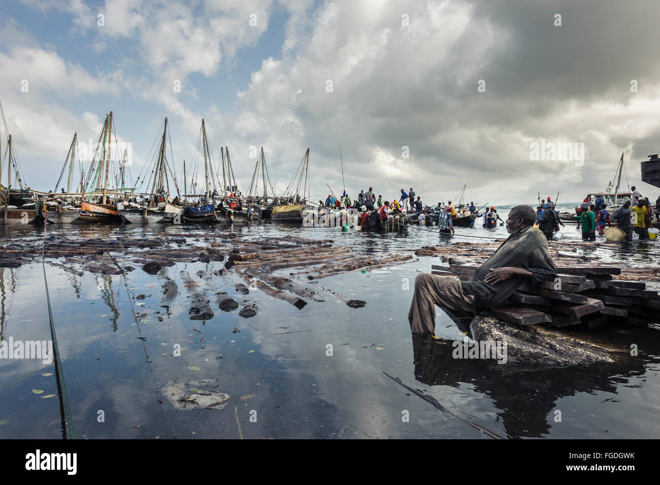 Fisherman resting with his feet in the water and looking at the crowd downloading goods from fishing boats. Stock Photo