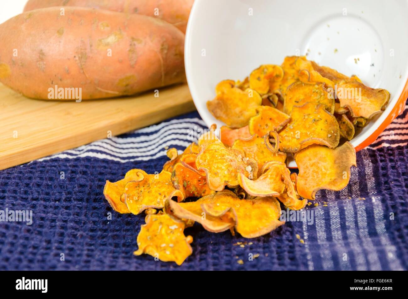 A bowl of homemade baked sweet potato chips poured over a kitchen towel and raw vegetable on a cutting board. A healthy sweetpot Stock Photo