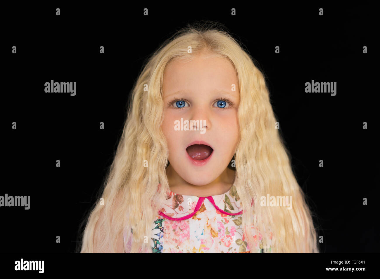 Young girl with long blonde hair with a surprised expression standing against a black background Stock Photo
