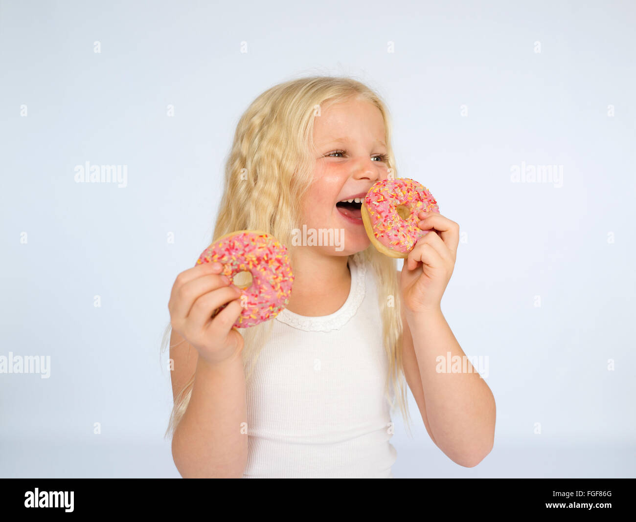 Young girl with long blonde hair holding two pink iced donuts, laughing Stock Photo