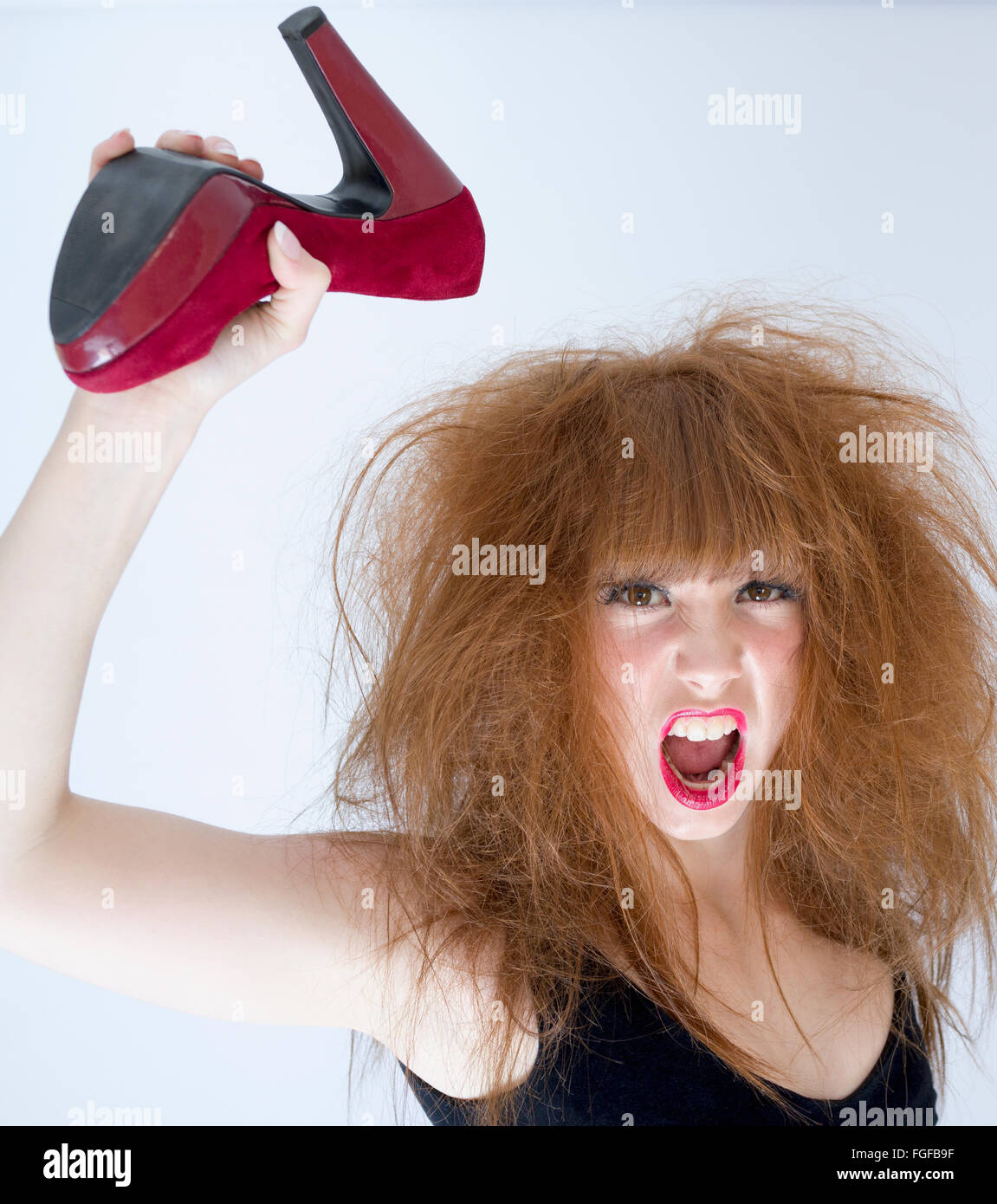 Woman with messy hair holding a red suede high heeled shoe in the air about to strike, screaming Stock Photo