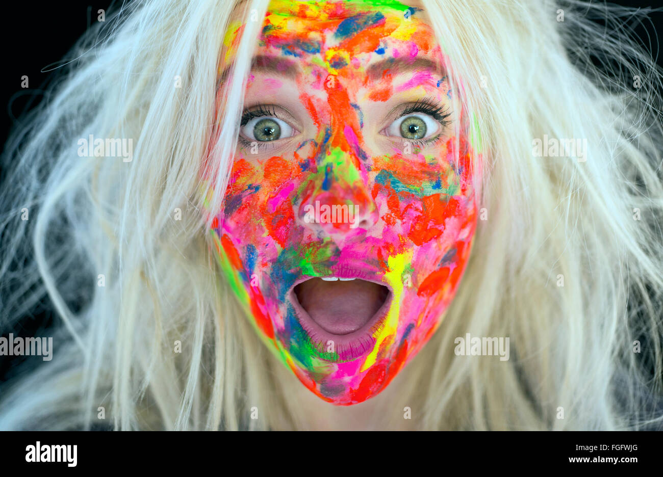 Woman with messy blonde hair and face covered in multi coloured paint with a surprised expression Stock Photo