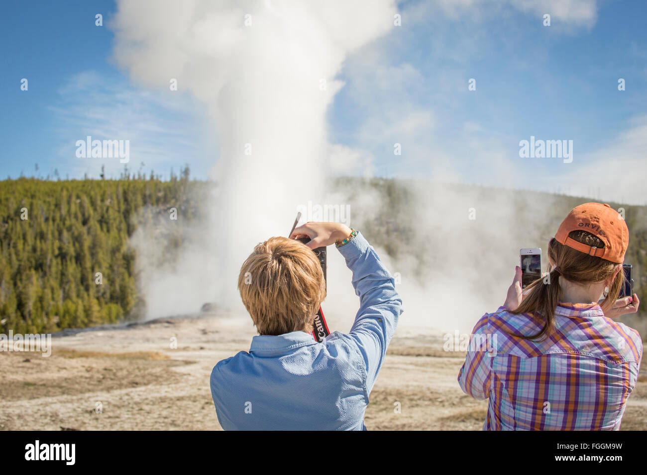 Two young women take cell phone photographs of Old Faithful. Stock Photo