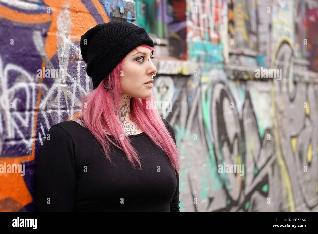 woman with pink hair piercings and tattoos leaning against graffiti wall Stock Photo
