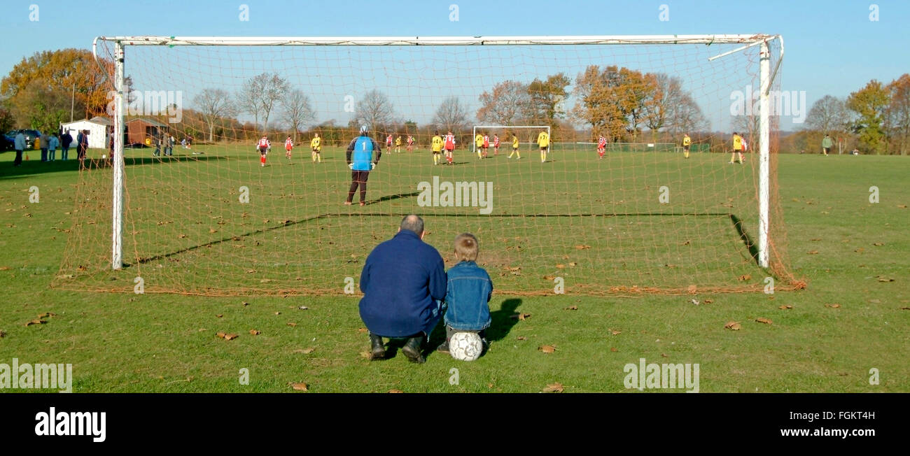 Father & son with ball behind goal post watching teen kids playing football match game village soccer pitch dad togetherness bonding with child UK Stock Photo