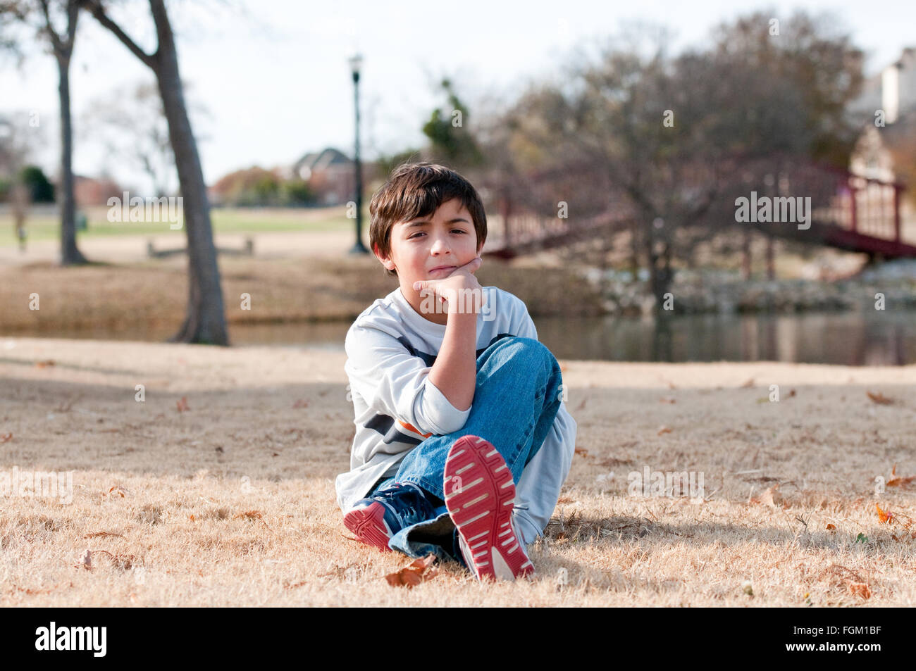 Cute little boy sitting in grass during the fall at a park with pond and bridge in background. Stock Photo