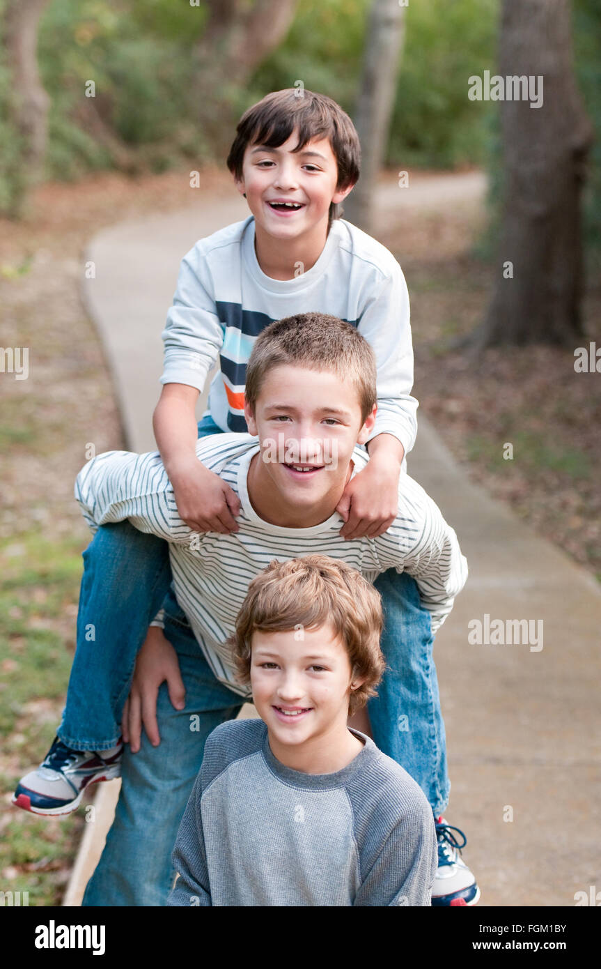 Group of three boys that are family and friends riding piggy back on sidewalk smiling at camera. Stock Photo