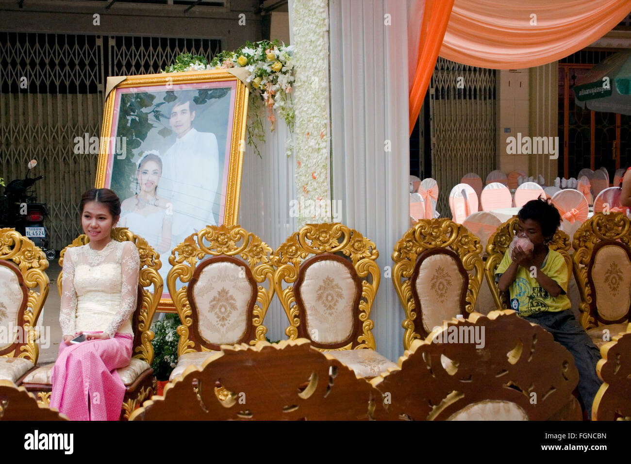 A young scavenger boy is sniffing glue while a bride sits near her wedding picture on a city street in Kampong Cham, Cambodia. Stock Photo