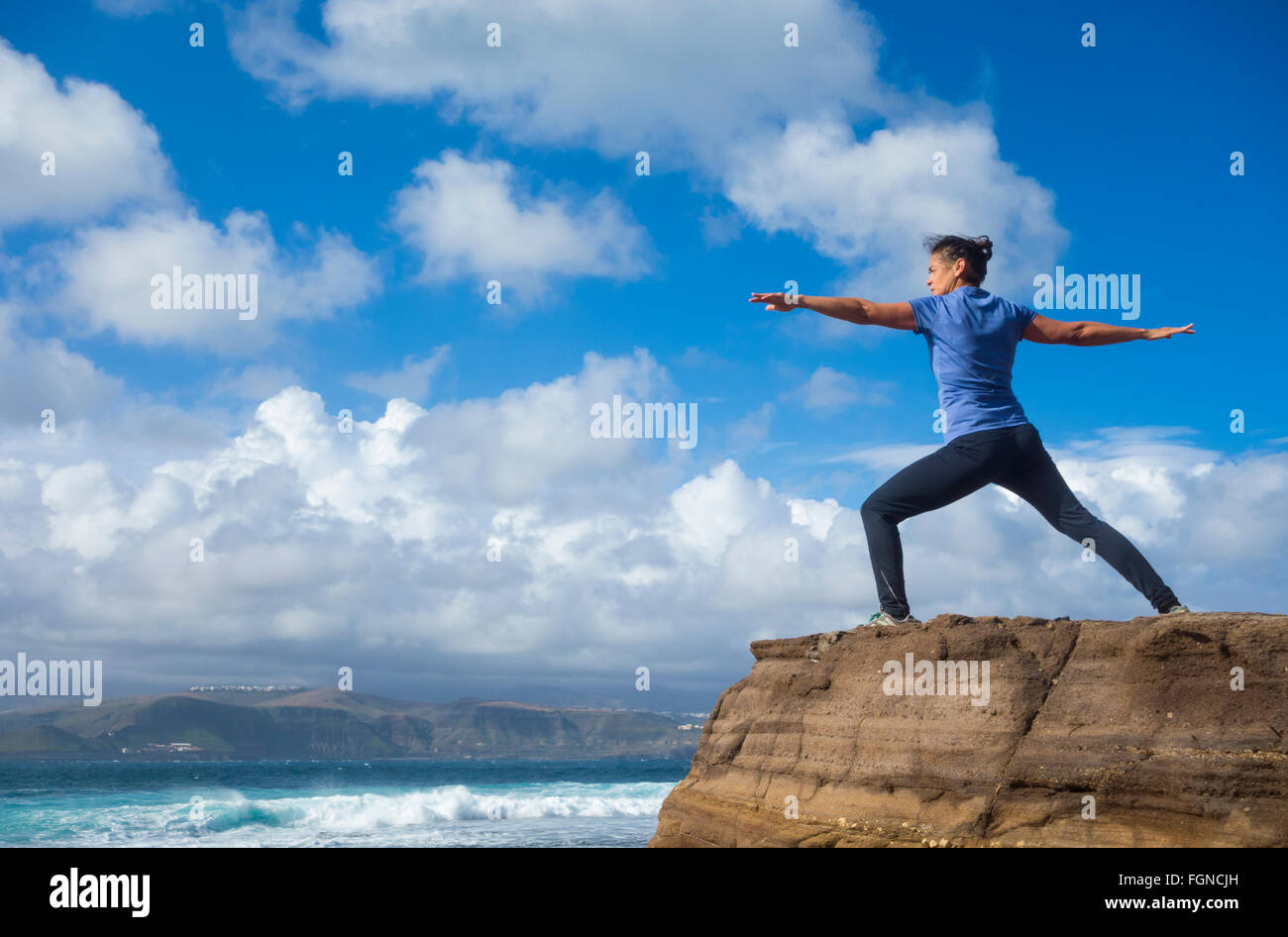 Mature female jogger warming up with Yoga stretching exercises on rock overlooking the sea. Stock Photo