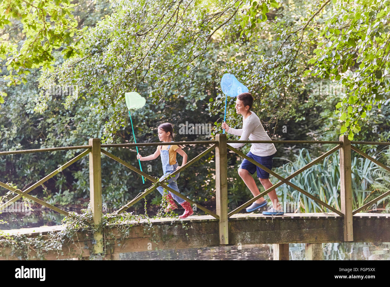 Brother and sister running with butterfly nets on footbridge in park Stock Photo