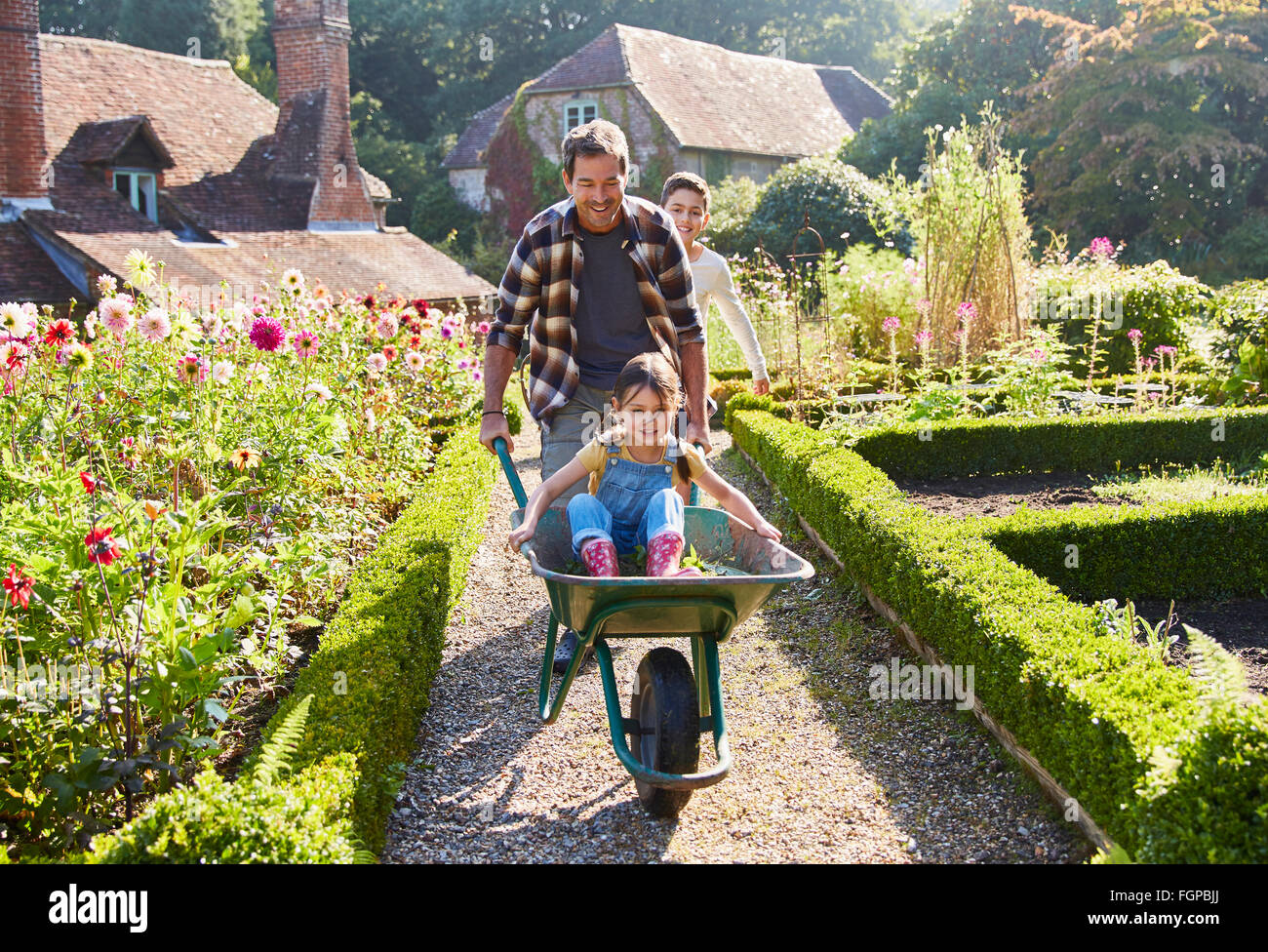 Father pushing daughter in wheelbarrow in sunny garden Stock Photo