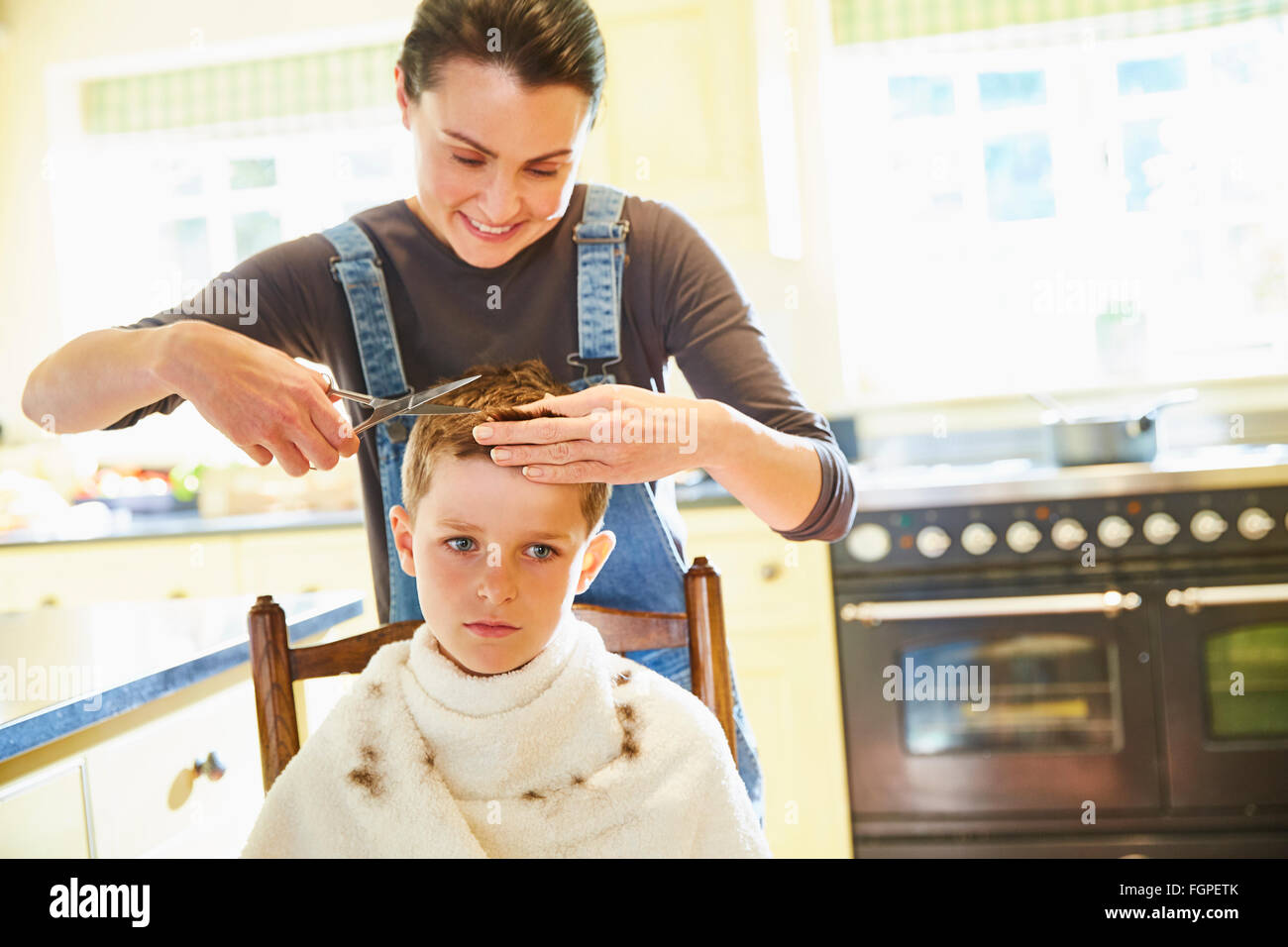 Unhappy boy getting haircut from mother in kitchen Stock Photo