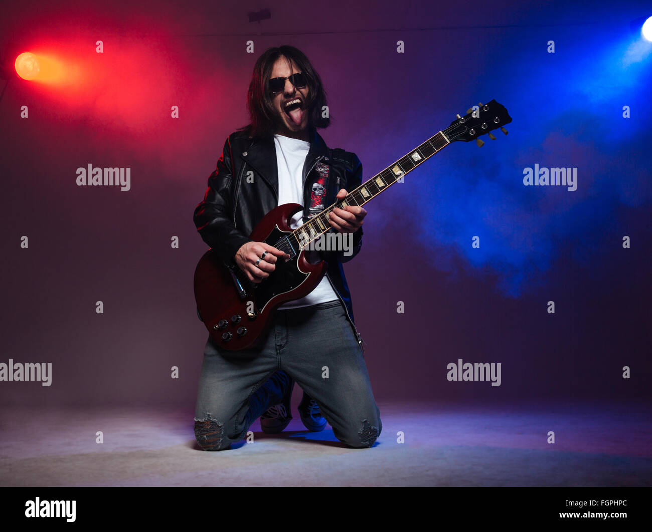 Crazy young guitarist with long hair playing electric guitar on his knees and shoing tongue over colorful smoky background Stock Photo