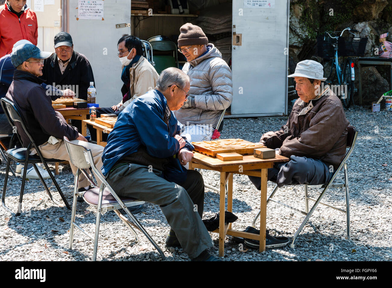 Japan, Kochi. Homeless unemployed men, all wearing coats, sitting at tables in the park playing shogi games in the springtime sunshine. Stock Photo