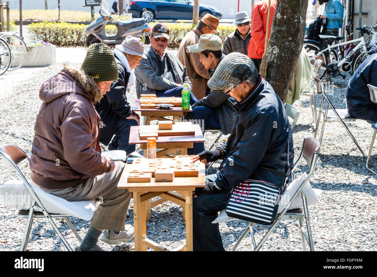Japan, Kochi. Homeless unemployed men, all wearing coats, sitting at tables in the park playing shogi games in the springtime sunshine. Stock Photo