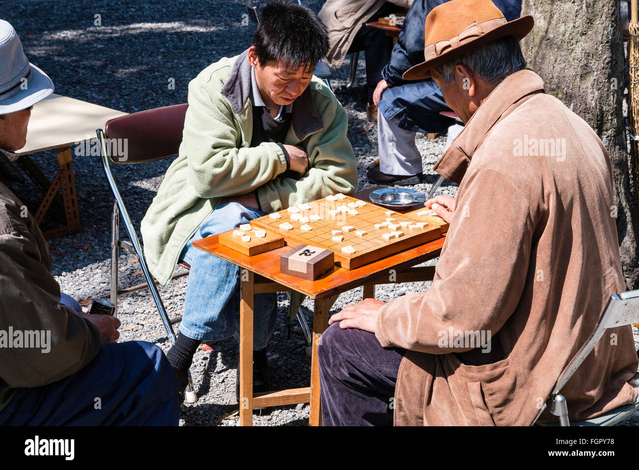 Japan, Kochi. Homeless unemployed men, all wearing coats, sitting at tables in the park playing shogi games in the springtime sunshine. Stock Photo