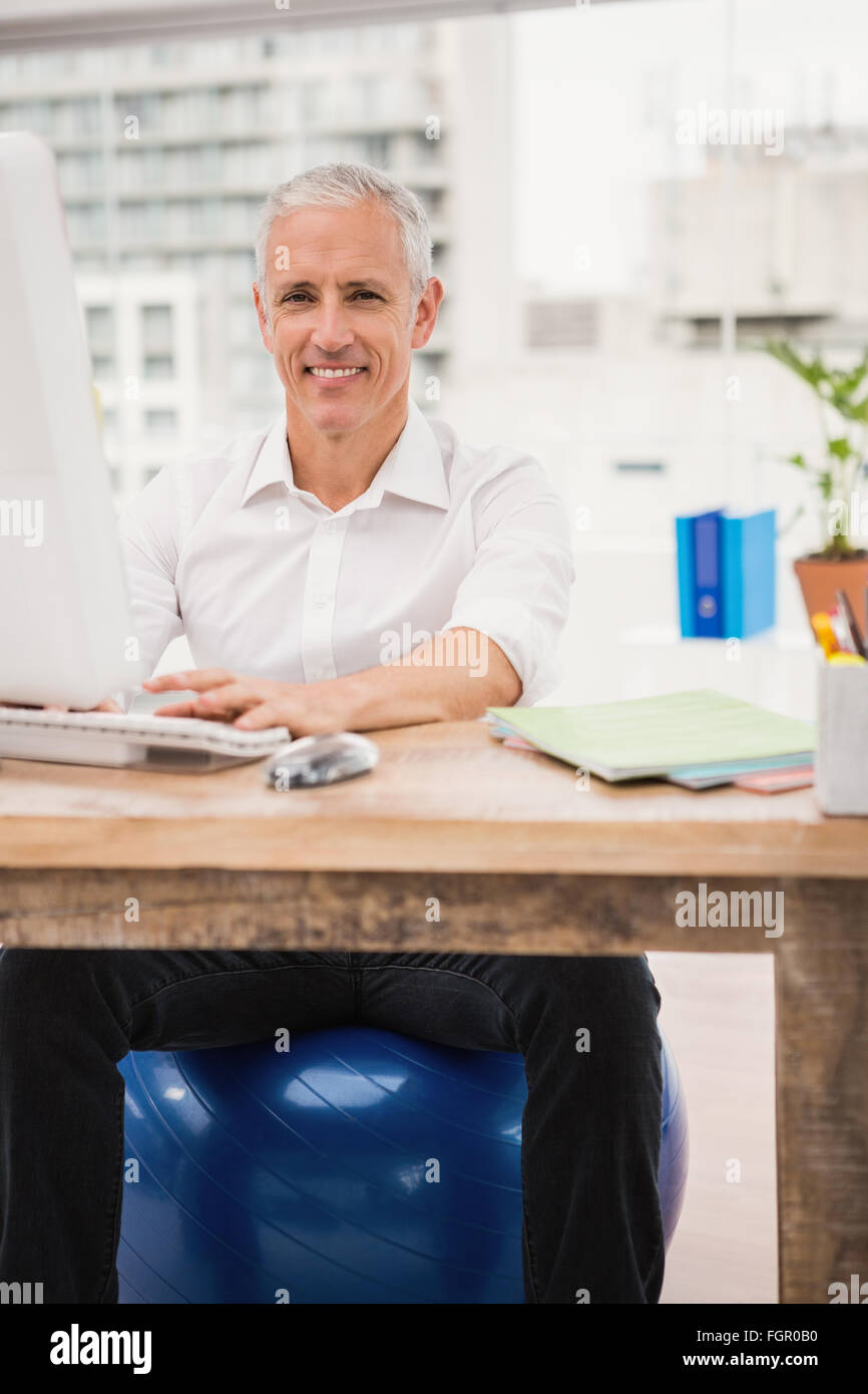 Smiling casual businessman sitting on exercise ball at desk Stock Photo