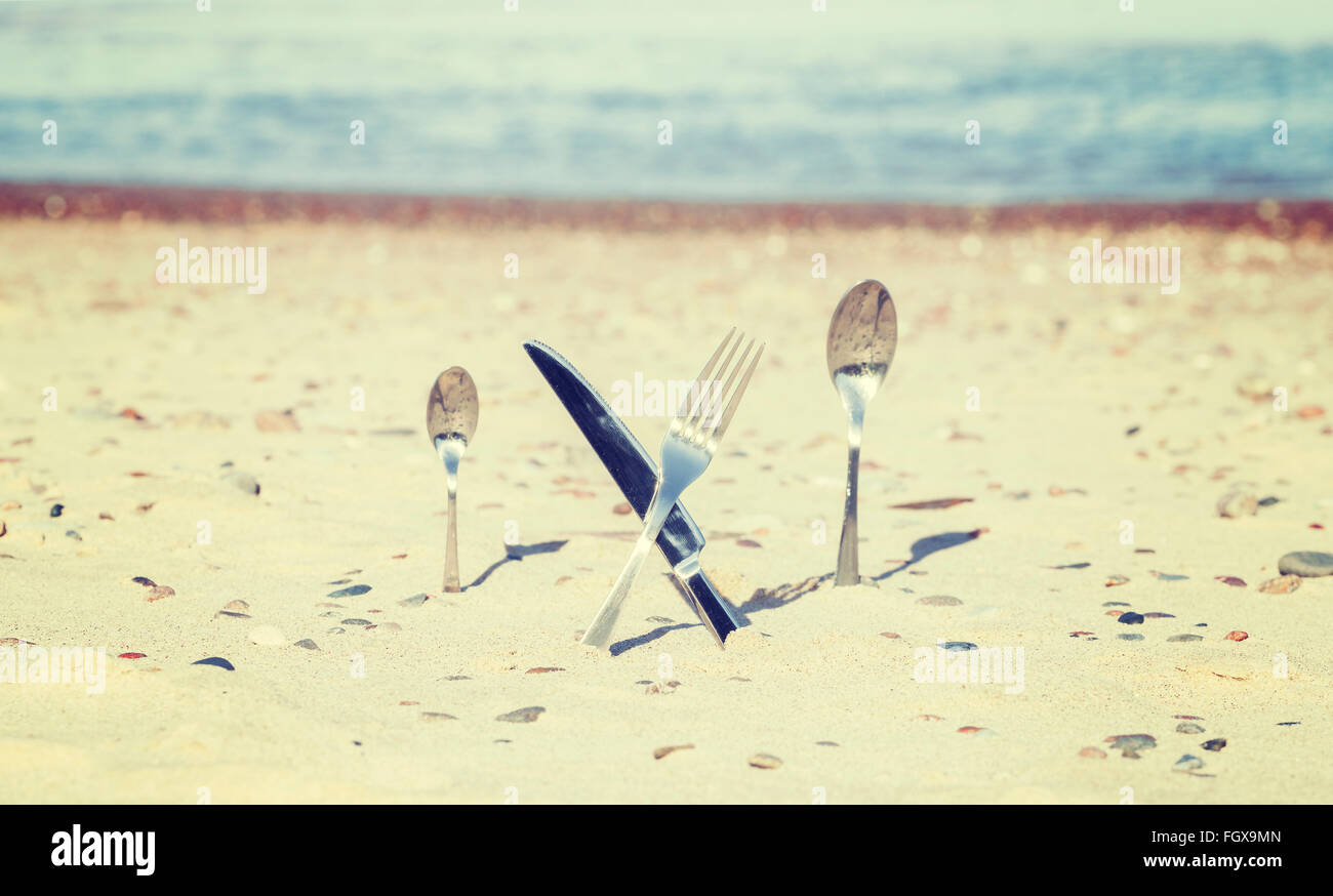 Vintage toned crossed knife and fork stuck in sand, shallow depth of field. Stock Photo