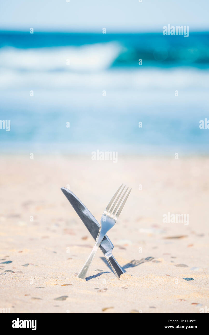 Crossed processed knife and fork stuck in sand, shallow depth of field. Stock Photo