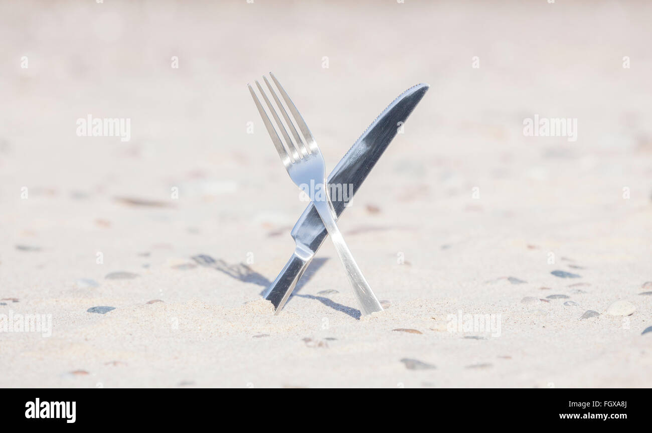 Crossed knife and fork stuck in sand, shallow depth of field. Stock Photo