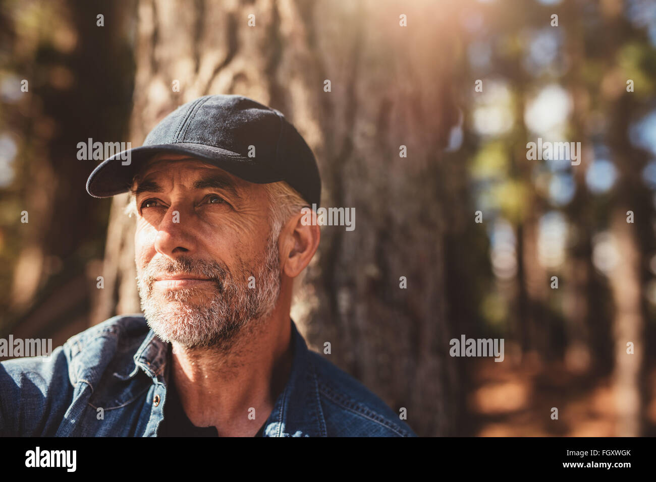 Close up portrait of senior man wearing cap looking away. Mature man with beard sitting in woods on a summer day. Stock Photo