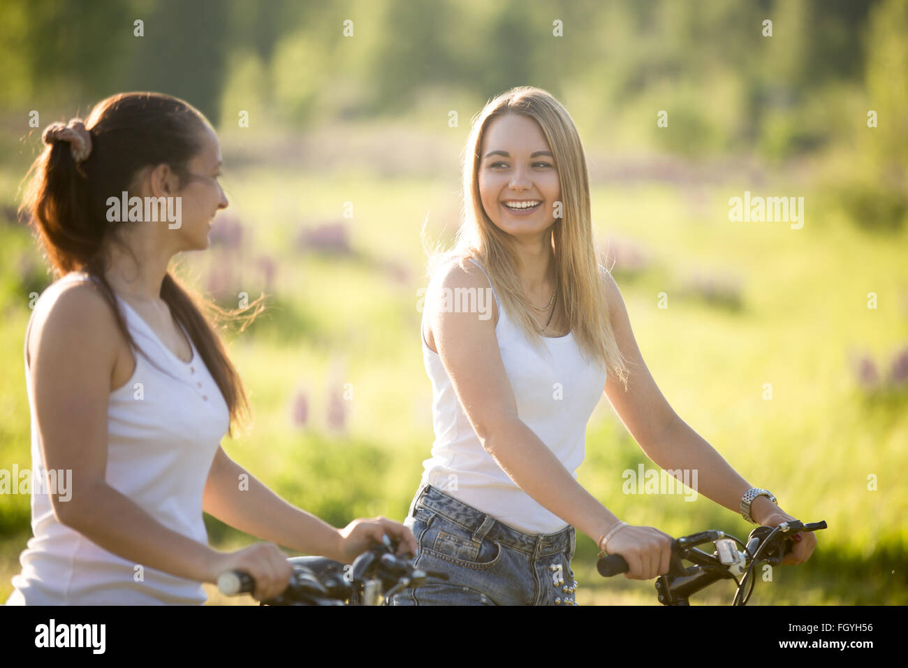 Two cheerful beautiful teenage girls wearing jeans shorts on bicycles in park on sunny summer day, having good time, laughing Stock Photo