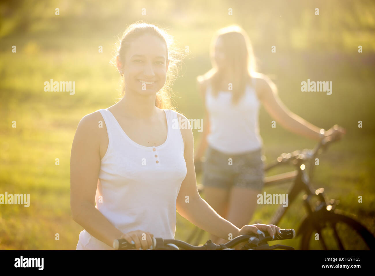 Two beautiful bicyclist girlfriends wearing casual white tank tops and jeans shorts standing with bikes in countryside park Stock Photo