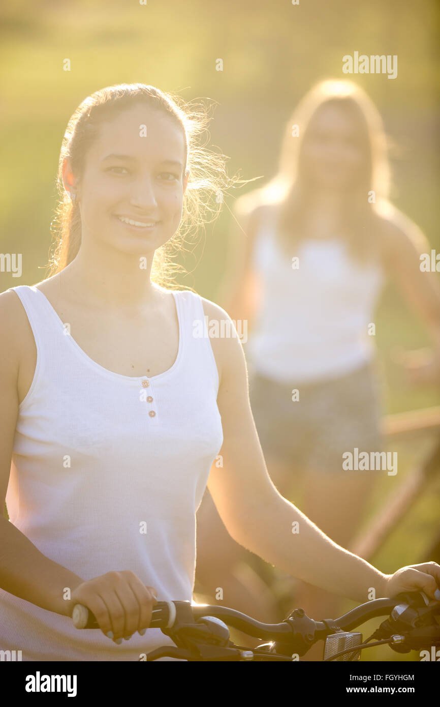 Two beautiful teenage girls with bikes wearing casual white tank tops and jeans shorts standing in countryside park on bright Stock Photo
