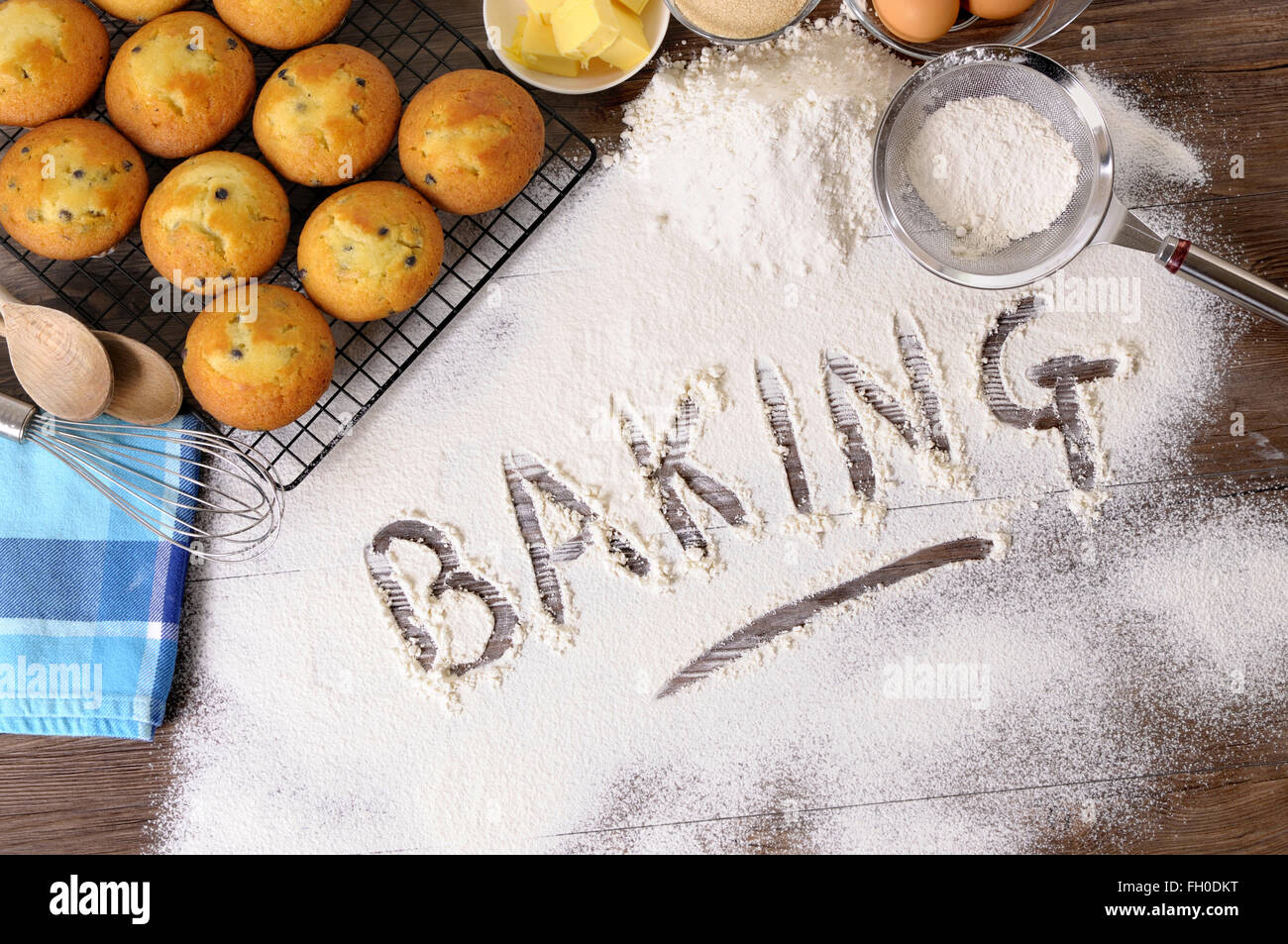The word Baking written in flour on a dark wood table with freshly baked muffins and ingredients. Stock Photo