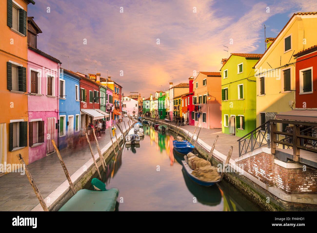 A view of colourful houses in Burano during the day along the canal. Colourful clouds can be seen in the sky. Stock Photo