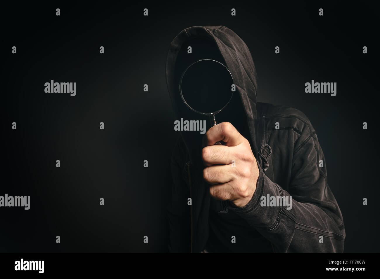 Spyware computer software, hooded spooky person with magnifying glass peeking at you Stock Photo