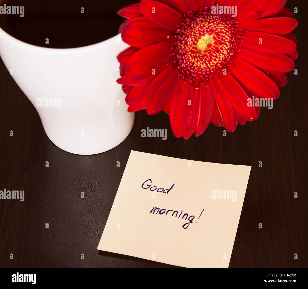 Red gerbera flower, cup of coffee and paper with text ''Good morning!' on the wooden desk. Selective focus, toned Stock Photo