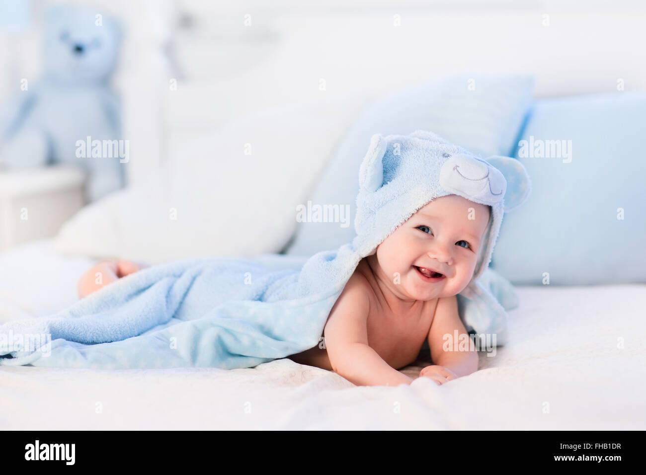 Baby boy wearing diaper and blue towel in white sunny bedroom. Newborn child relaxing in bed after bath or shower. Stock Photo