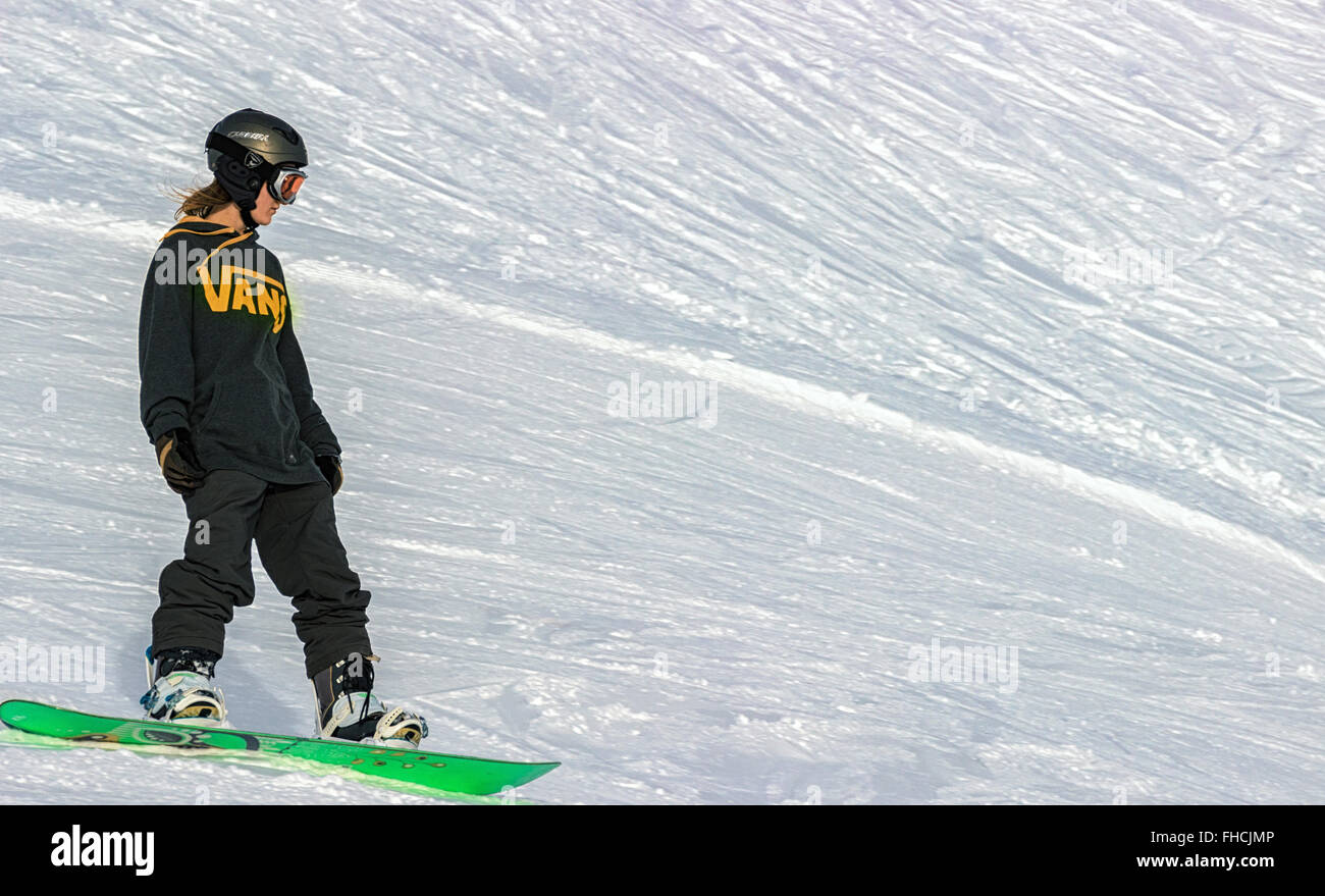 A cool young woman in a black sports uniform, helmet and snow goggles sliding down on a green snowboard at a sunny day. Stock Photo