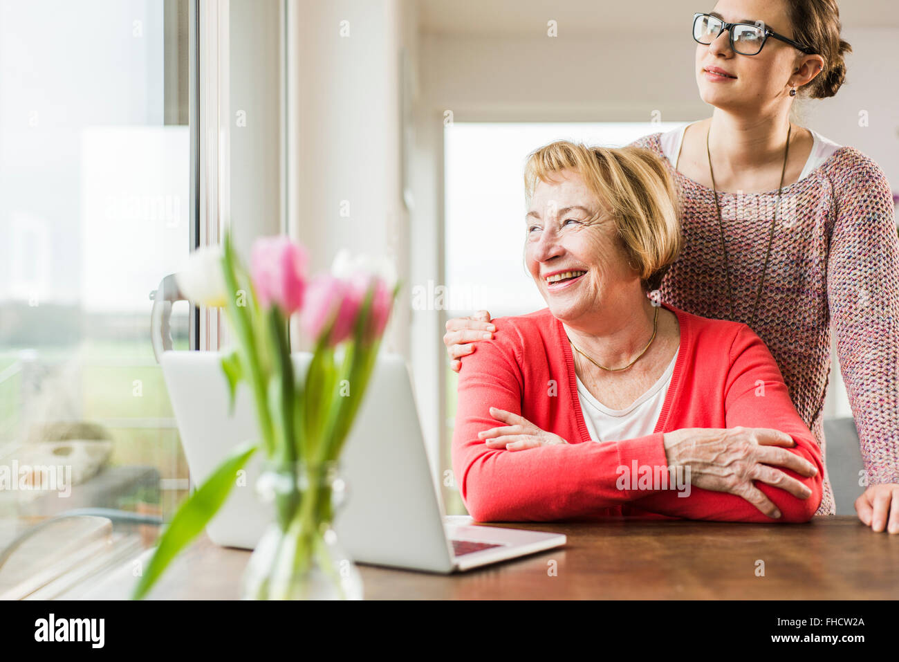 Smiling young woman with senior woman at table with laptop looking out of window Stock Photo