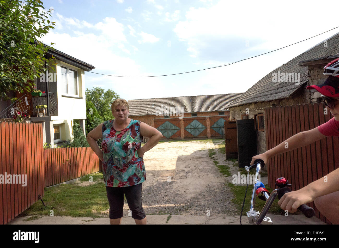 Woman with hands on hips stands guard as bicyclers pass by her driveway. Rzeczyca Central Poland Stock Photo