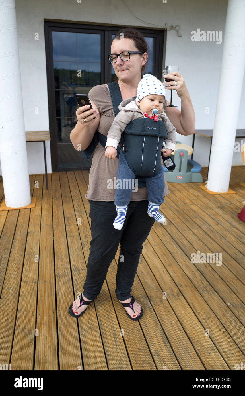 Multitasking mom with wine in one hand and cell phone in the other carrying Jack in a Babybjorn carrier. Zawady Central Poland Stock Photo
