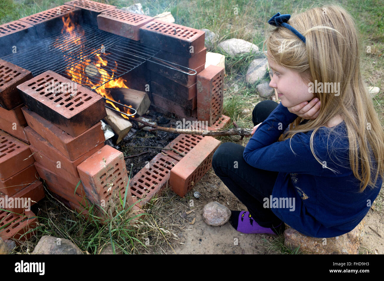 Young Polish girl age 8 musing while poking fire with a stick in outside grill fireplace. Zawady Central Poland Stock Photo