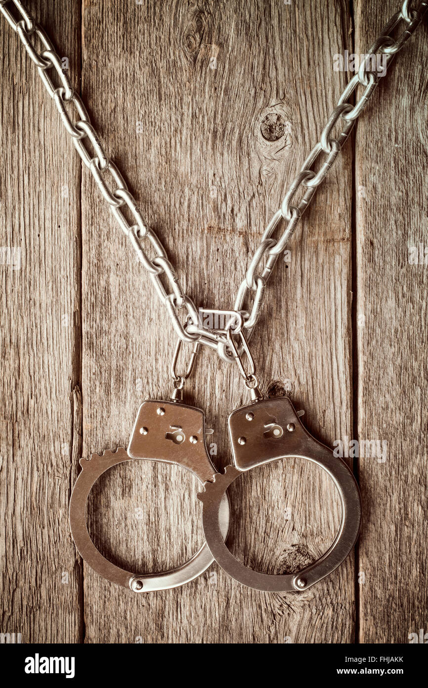Handcuffs hanging on the chain against old wooden wall. Stock Photo