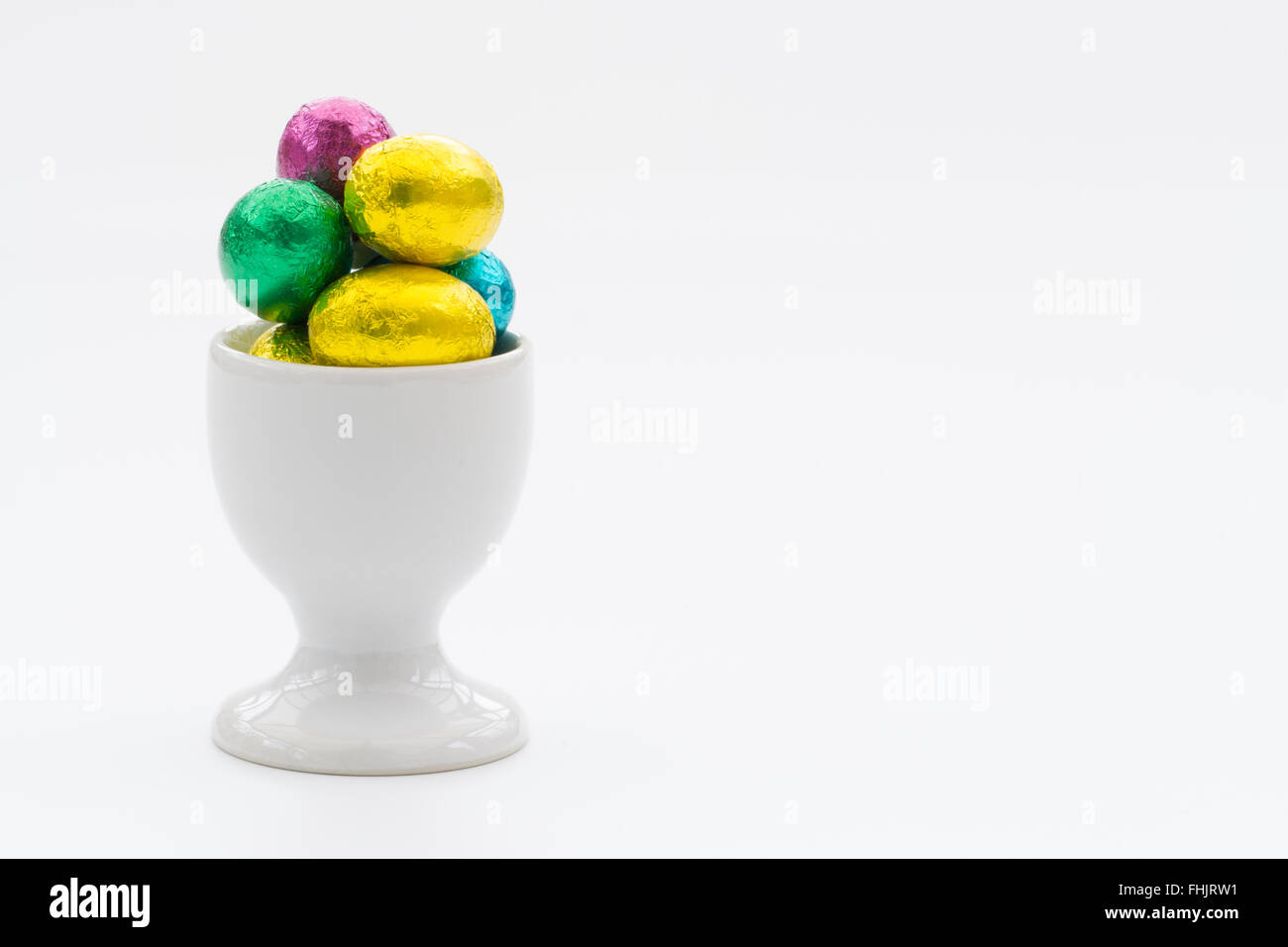 A white egg cup filled with small, chocolate Easter eggs on an isolated white background. Stock Photo