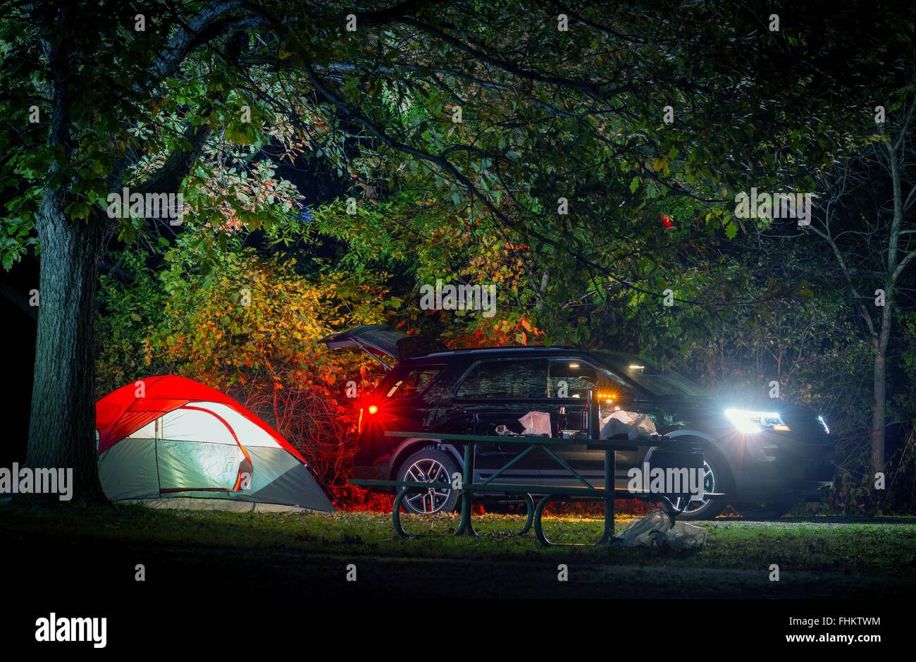 Summer Overnight Tent Camping. Modern Sport Utility Vehicle and Illuminated Small Double Tent Under Old Tree. Campsite on Camp Stock Photo