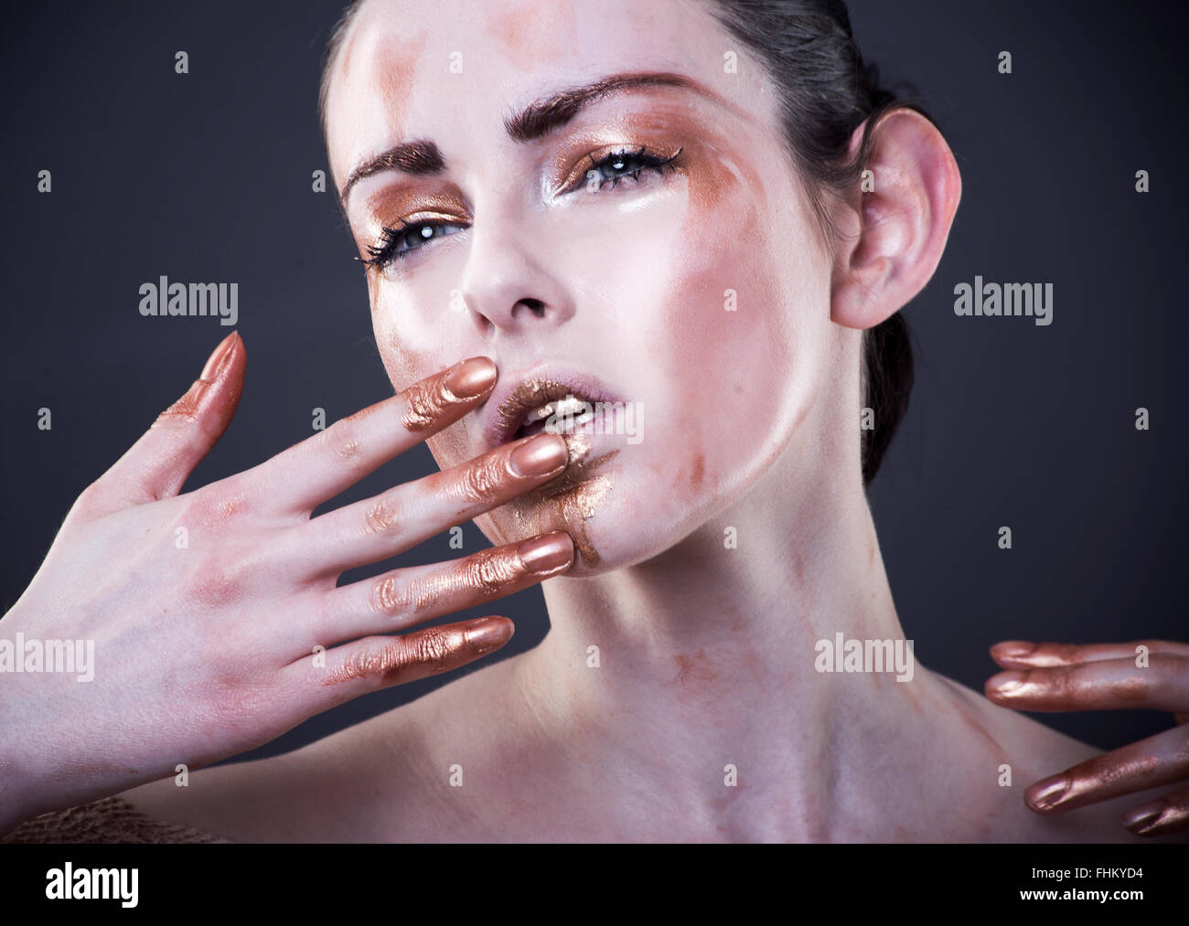 Beautiful fashion model with hand near mouth, brass paint make up on fingers and face, eyes slightly closed, intense expression Stock Photo