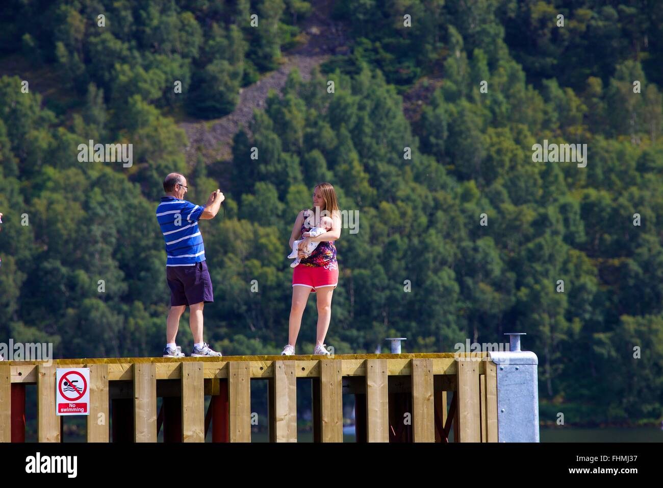 Lake District National Park. Man taking photograph of his wife and baby with smart phone on a jetty in lake Ullswater. Stock Photo