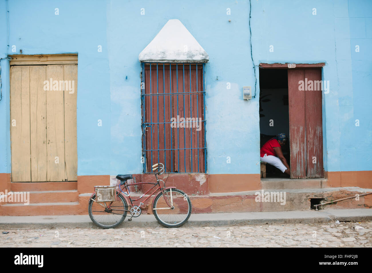 A bicycle parked in front of some doors in a blue building in a street of Trinidad, Cuba. A woman sits in one of the doorways. Stock Photo